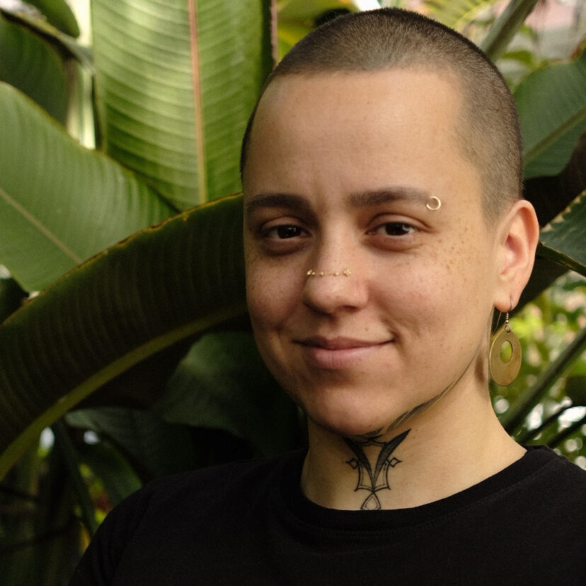 Nellie Grant, LMT A photo of Nellie Grant, the owner and sole practitioner at Cloudbelly Bodywork LLC. The photo shows their smiling face turned toward the camera with warmth and comfort in their brown eyes. They are wearing wire-framed glasses, dangly earrings, and a their